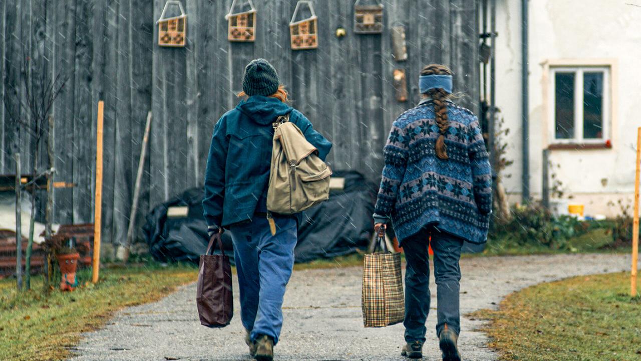 © ORF/Wega Film Marian und Gerti gehen bei leichtem Regen mit Taschen über einen Weg zu einem alten Hofgebäude.