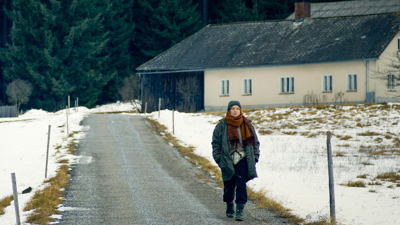 © ORF/Wega Film Miriam geht im Winter in einer schneebedeckten Landschaft über einen schmalen Weg an einem Hof vorbei.