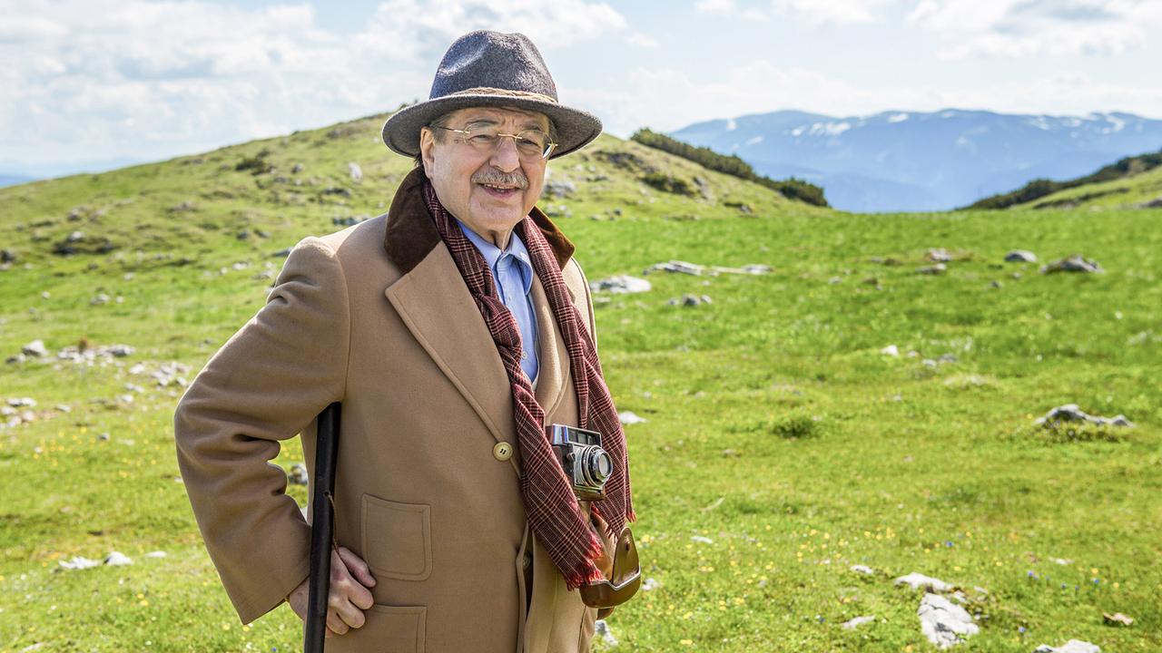 Erwin Steinhauer mit Hut, Wanderstock und Kamera steht auf einer grünen Alm am Schneebergvor einer Bergkulisse.