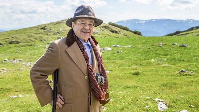© ORF/FOR TV/Peter Aigner Erwin Steinhauer mit Hut, Wanderstock und Kamera steht auf einer grünen Alm am Schneebergvor einer Bergkulisse.