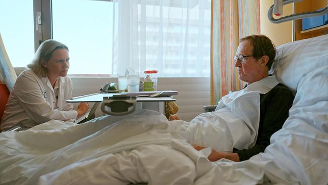 © ORF/Metafilm GmbH/Klemens Koscher Primarärztin Eva Katharina Masel sitzt an einem Krankenhausbett und spricht mit Patient Matthias Frisch, der im Bett liegt.