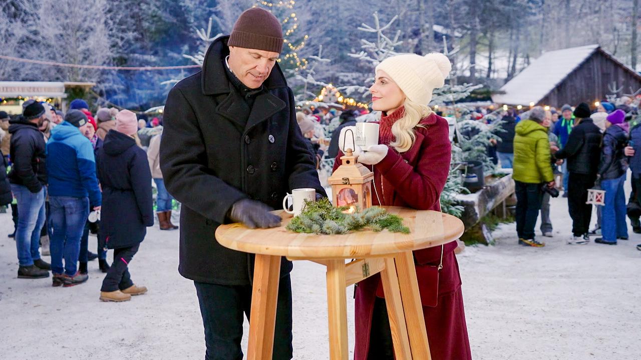 Armin Assinger und Silvia Schneider trinken heiße Getränke an einem Holzstehtisch auf einem belebten und winterlich-verschneiten Weihnachtsmarkt im Freien.