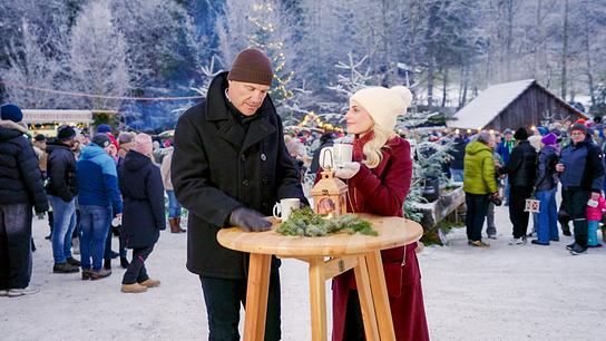 © ORF Armin Assinger und Silvia Schneider trinken heiße Getränke an einem Holzstehtisch auf einem belebten und winterlich-verschneiten Weihnachtsmarkt im Freien.