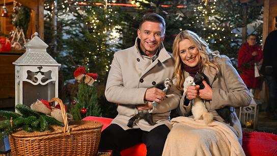 Marco Ventre und Stefanie Hertel in winterlicher Kleidung sitzen auf einer Bank vor Weihnachtsbäumen und kleine Tiere auf dem Schoß.