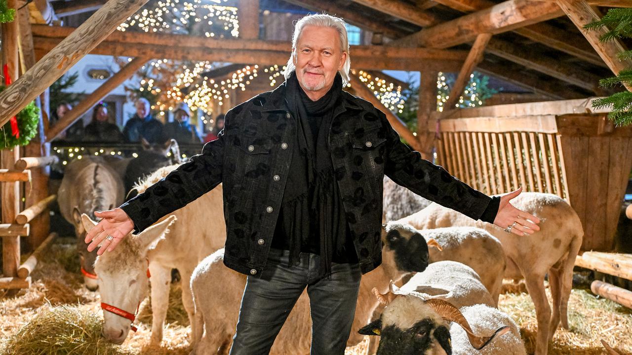 Johnny Logan steht mit ausgebreiteten Armen vor einer Gruppe von Eseln und Schafen in einem weihnachtlich beleuchteten Stall.