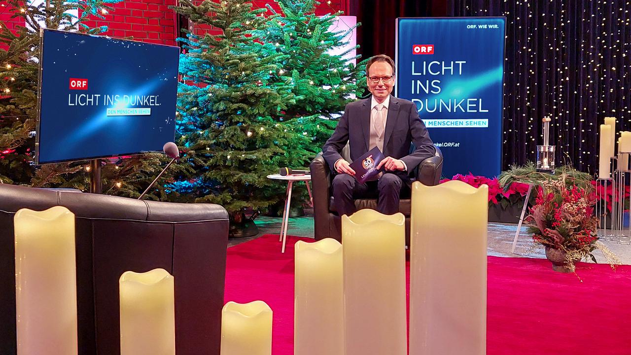 Günther Madlberger im festlich dekorierten TV-Studio auf der ORF‑„Licht ins Dunkel“-Bühne, umgeben von Kerzen, Weihnachtsbäumen und Studiobeleuchtung.