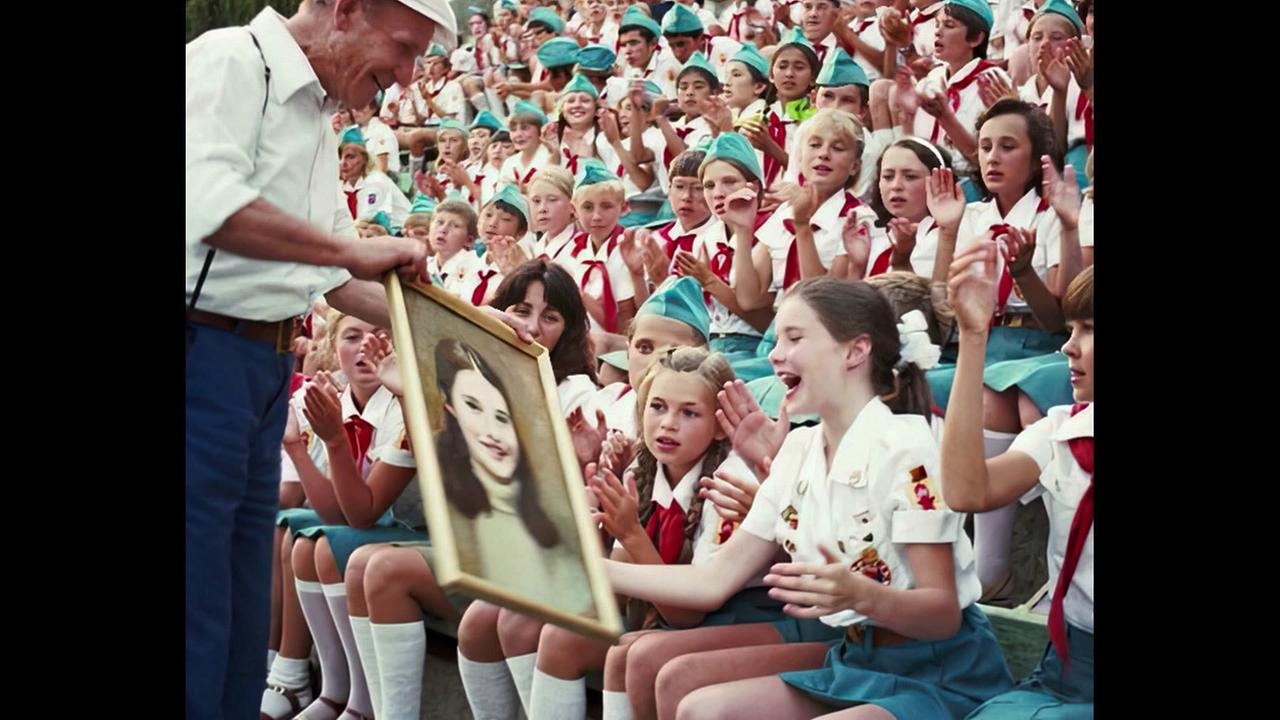 Eine große Gruppe uniformierter Kinder und Jugendlicher sitzen bei einem Jugendcamp in der UdSSR auf Tribünen und klatschen, während ein Mann der zehnjährigen Samantha Smith ein gerahmtes Bild überreicht. 