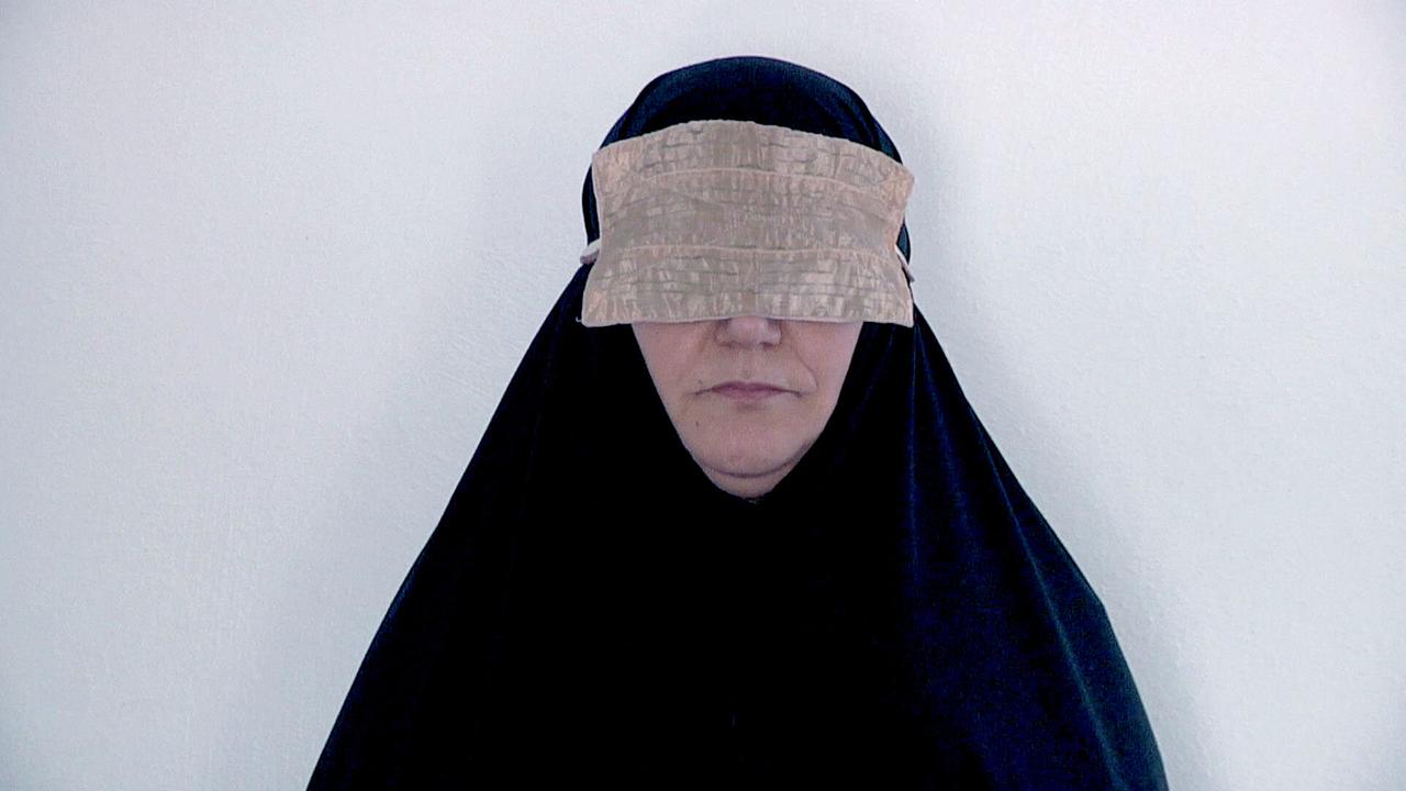 Eine Frau mit einer Augenbinde ist mit einem Tschador verhüllt, einem traditionellen, iranischen Umhang vor einem schlichten, hellen Hintergrund.