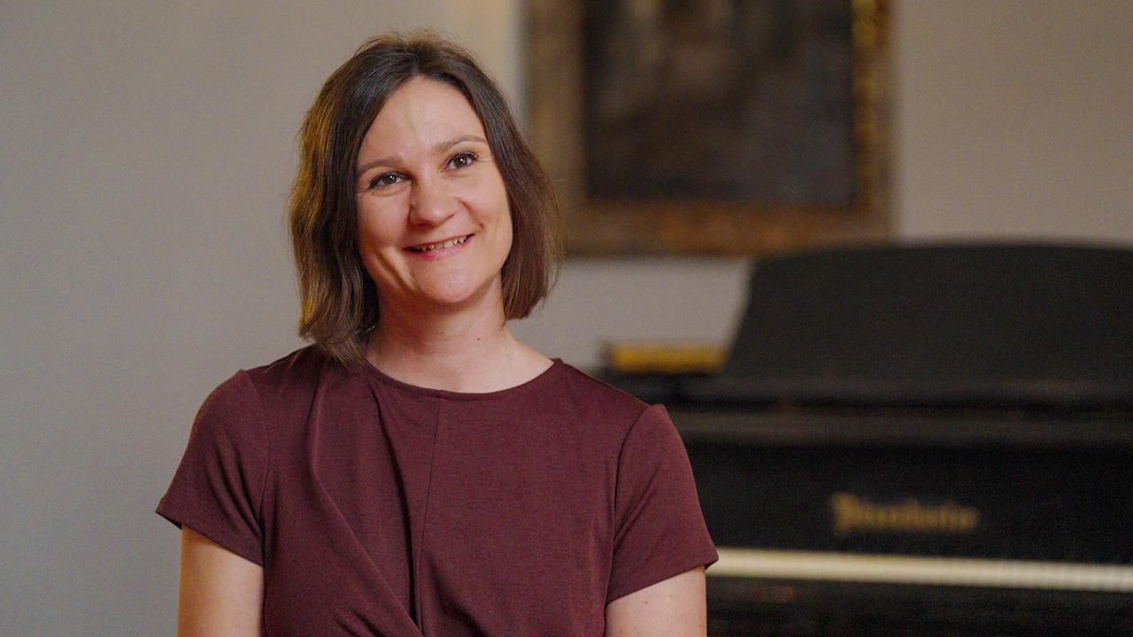© ORF/Raumfilm Dommusikkapellmeisterin Melissa Dermastia mit schulterlangem Haar in einem bordeauxfarbenen Oberteil sitzt bei einem Interview in einem Raum vor einem Klavier.