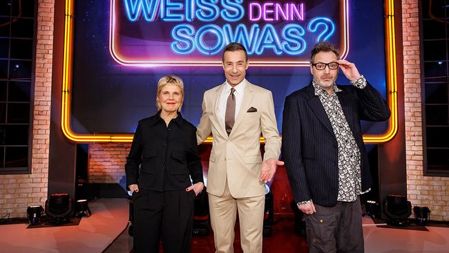 Cordula Stratmann, Kai Pflaume und Paul Panzer stehen im TV-Studio vor einem leuchtenden Neon‑Logo mit der Aufschrift "Wer weiß denn sowas?"