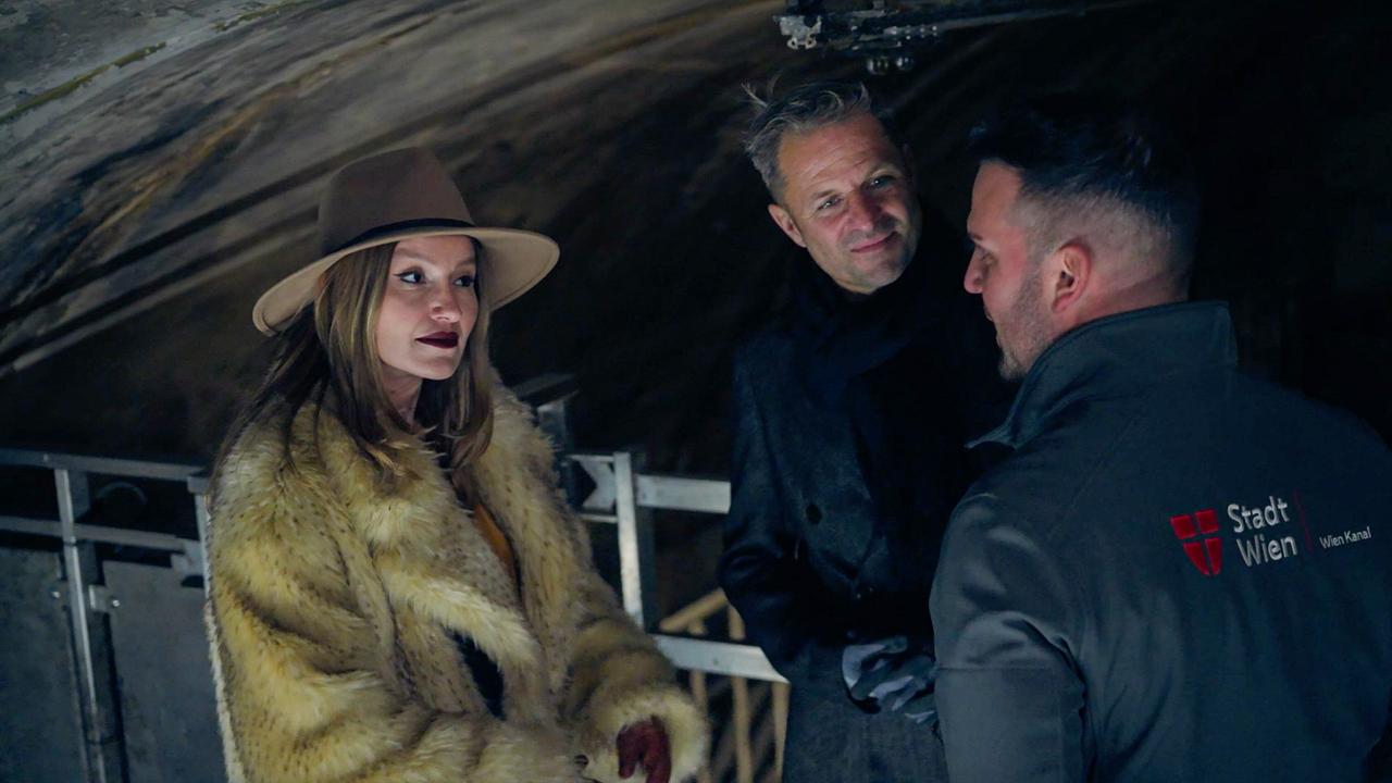 Malarina und Philipp Hochmair in Winterkleidung stehen mit Leo Kaps von der Stadt Wien in einem Tunnel auf einer Metallplattform und sprechen miteinander.