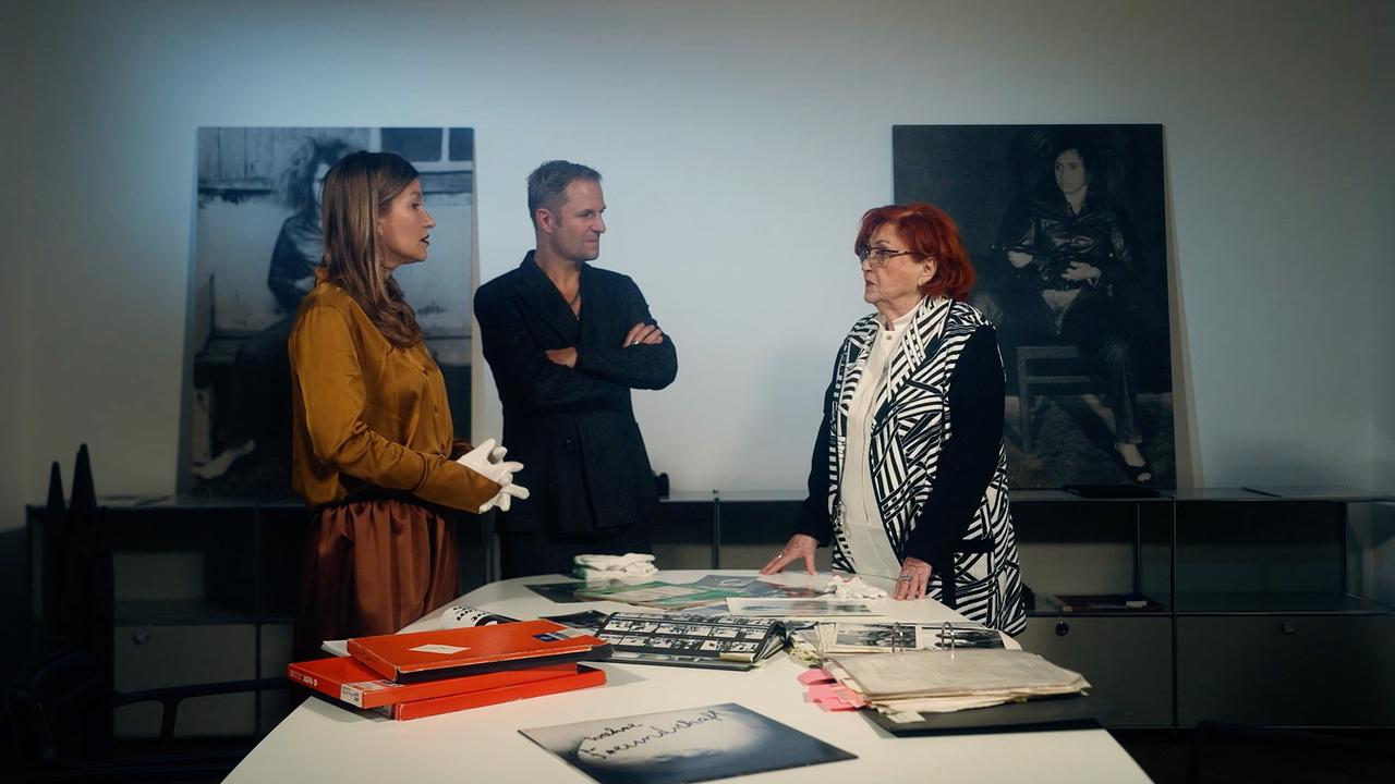 Malarina und Philipp Hochmair zu Besuch bei der Künstlerin Valie Export: Sie stehen in einem Raum an einem Tisch mit Fotos und Dokumenten vor großformatigen Schwarzweiß-Bildern.