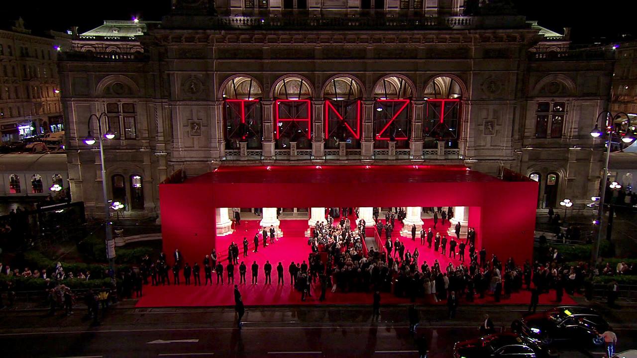 Eine nächtliche Totalansicht der Wiener Staatsoper zeigt einen großen roten Eingangsbereich mit rotem Teppich, umgeben von zahlreichen Gästen und Sicherheitskräften.