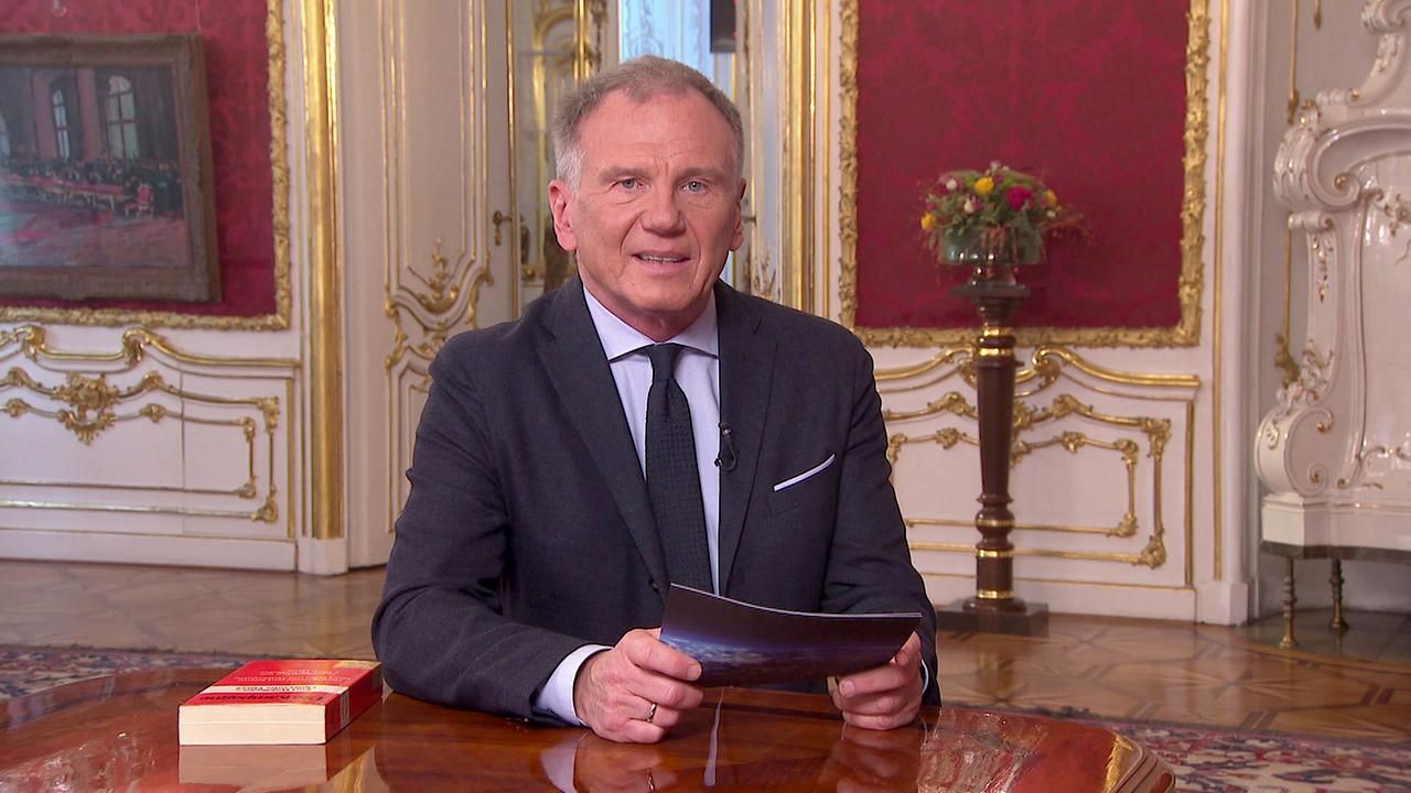 © ORF Armin Wolf sitzt an einem Tisch in einem prunkvollen Raum mit goldverzierten Wänden und hält ein Blatt Papier in den Händen.