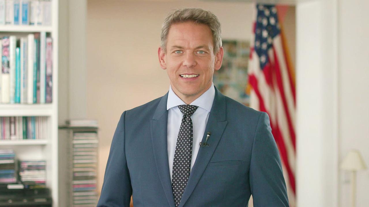 © ORF/Hannes Drapal Martin Thür im Anzug steht in einem hellen Büro vor einem Bücherregal, im Hintergrund ist eine US‑amerikanische Flagge zu sehen.