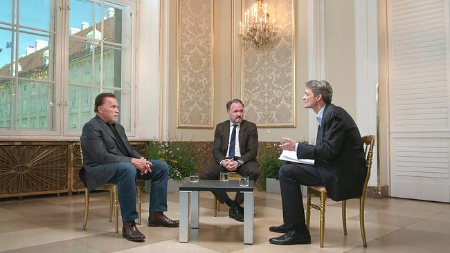 © ORF/Roman Bagner Arnold Schwarzenegger sitzt mit zwei weiteren Männern in einem eleganten Raum auf Stühlen im Gespräch an einem kleinen Tisch.