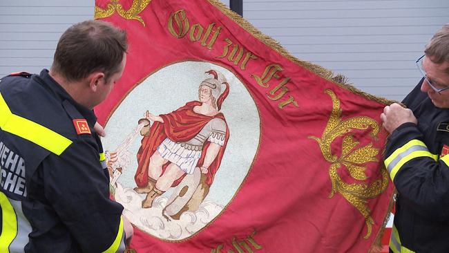 Zwei Feuerwehrleute halten eine Fahne mit der Abbildung des Heiligen Florians.