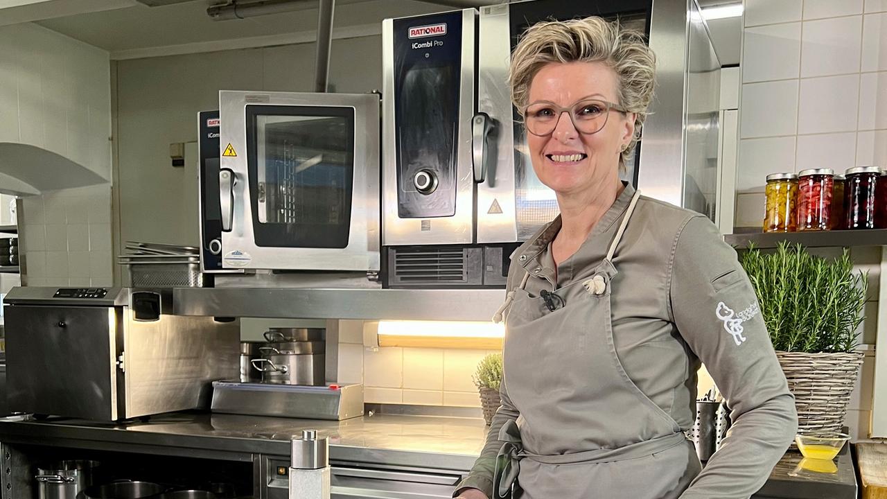 Renate Schaufler steht in ihrer Küche, Geräte und ein Topf mit Kräutern im Hintergrund.