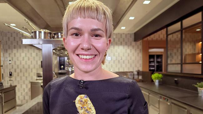 © ORF/Ab Hof TV Produktion Köchin Janine Wieland in Kochjacke hält ein Mädesüßparfait am Stiel mit sichtbaren Blüten in einer modernen Küche.