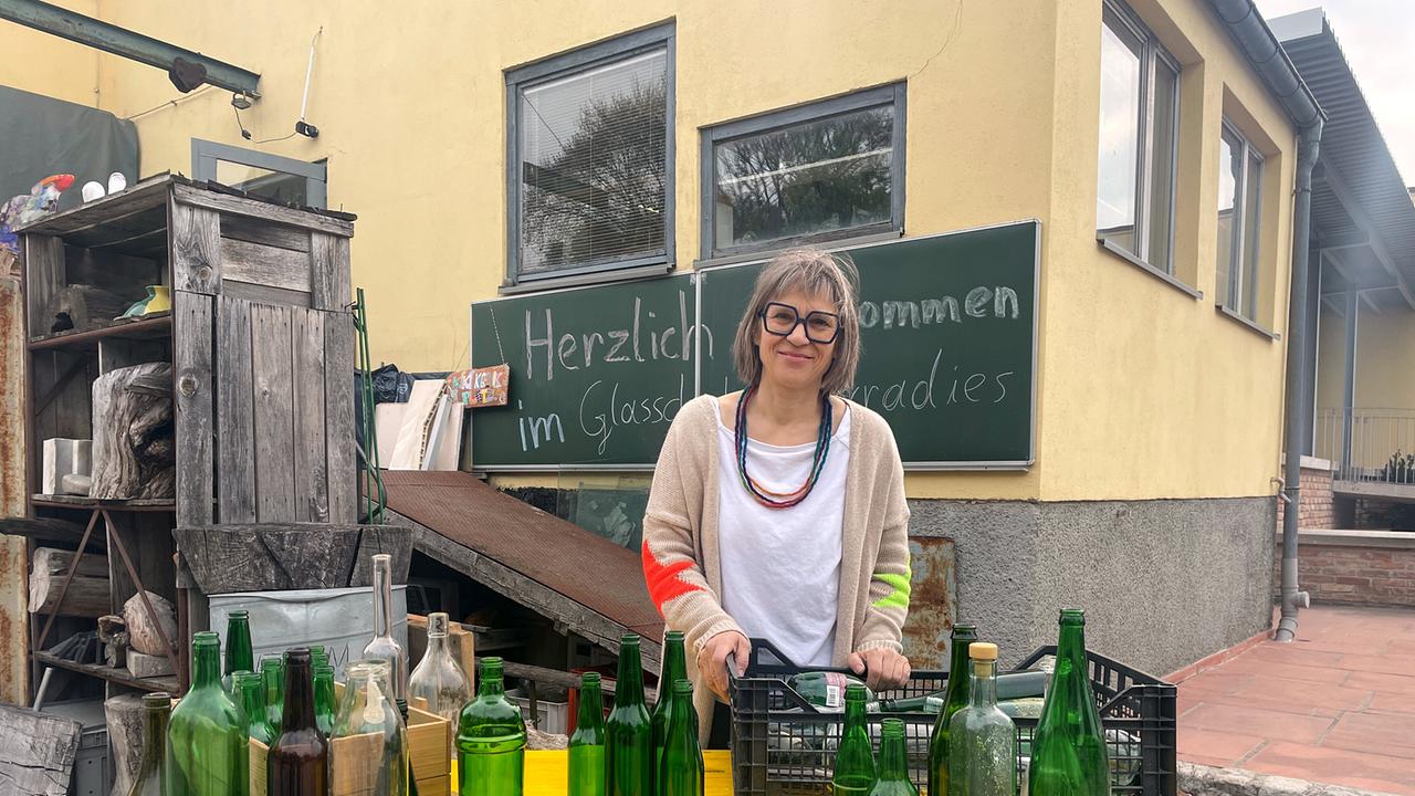 © ORF/Ab Hof TV Produktion Glaskünstlerin Simone Svoboda hinter einem Tisch im Freien mit leeren Glasflaschen.