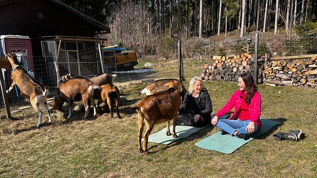Christina Goritschnig und Maggie Entenfellner sitzen auf Yogamatten im Freien in der Sonne, umgeben von mehreren Ziegen.