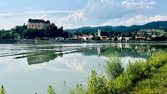 © ORF/Ab Hof TV Produktion Die Stadt Grein an der Donau mit Schloss Greinburg auf einem Hügel, Kirche und Häusern am Flussufer, spiegelnde Donau.