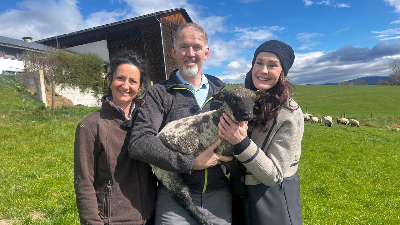 © ORF/Ab Hof TV Produktion Daniela und Wolfgang Ochabauer und Maggie Entenfellner stehen auf einer Wiese, Wolfgang Ochabauer hält ein Lamm im Arm.