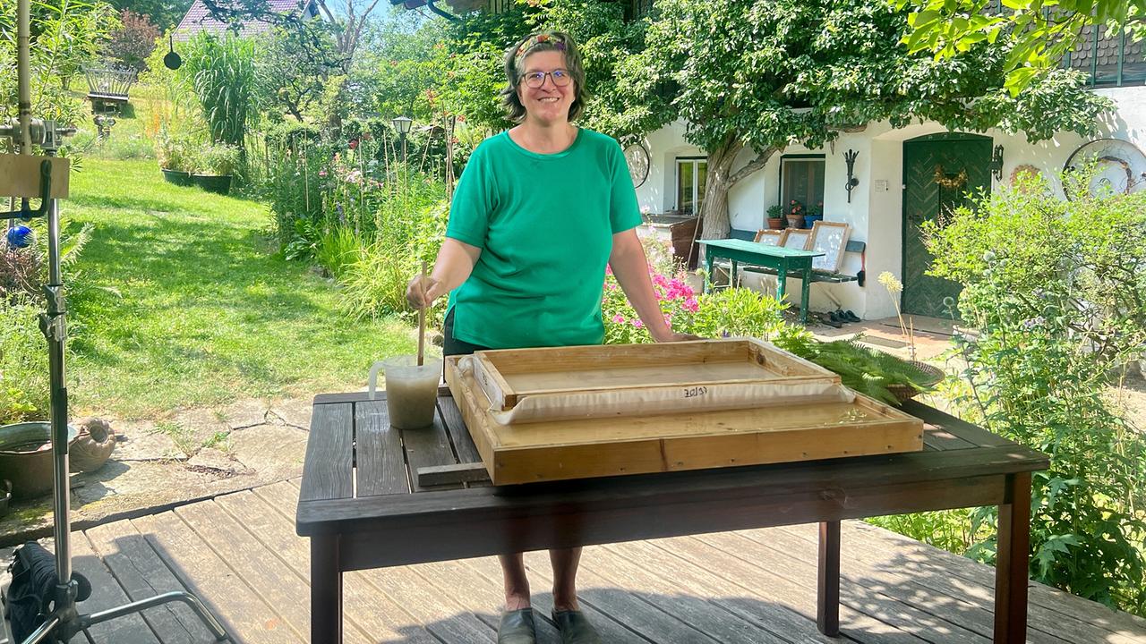 Michaela Metzler steht lächelnd mit Papierschöpfrahmen am Holztisch im Garten vor einem pflanzenreichen Haus.