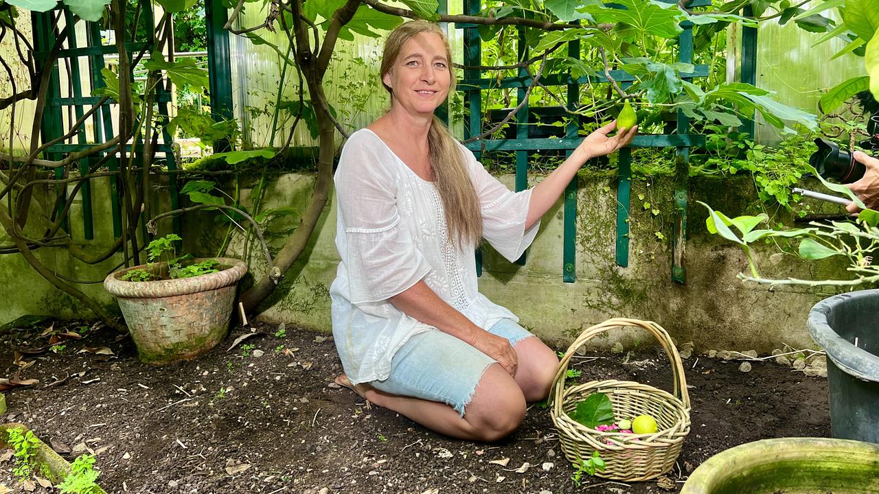 Elisabeth Mayer kniet in ihrem Naturgarten neben einem Korb voller Früchte und hält eine Feige in den Händen.