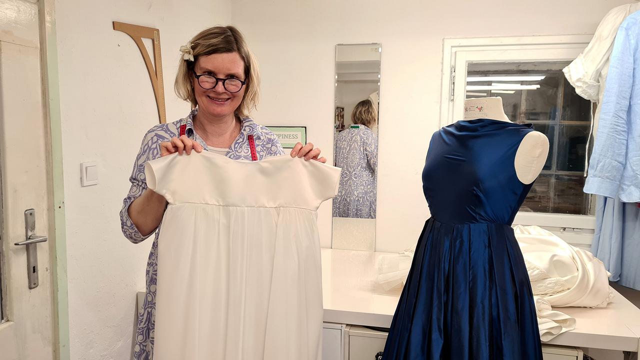 Schneidermeisterin Reingard Ruzicka hält ein Taufkleid in Händen, neben einer Schneiderpuppe stehend.