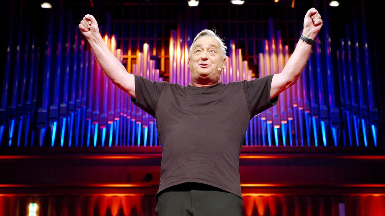 Andreas Vitásek steht auf einer Bühne vor einer großen, farbig beleuchteten Orgelwand und hebt beide Arme triumphierend nach oben.