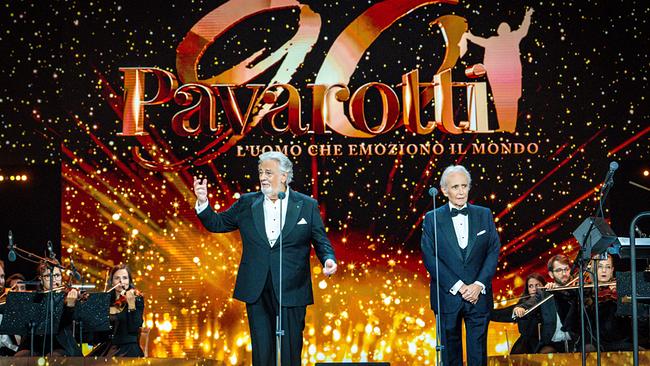 © ORF/C Major Entertainment/Francesco Prandoni Plácido Domingo und José Carreras stehen auf einer festlich beleuchteten Konzertbühne vor einem Orchester und dem großen Schriftzug „Pavarotti 90“ im Hintergrund.