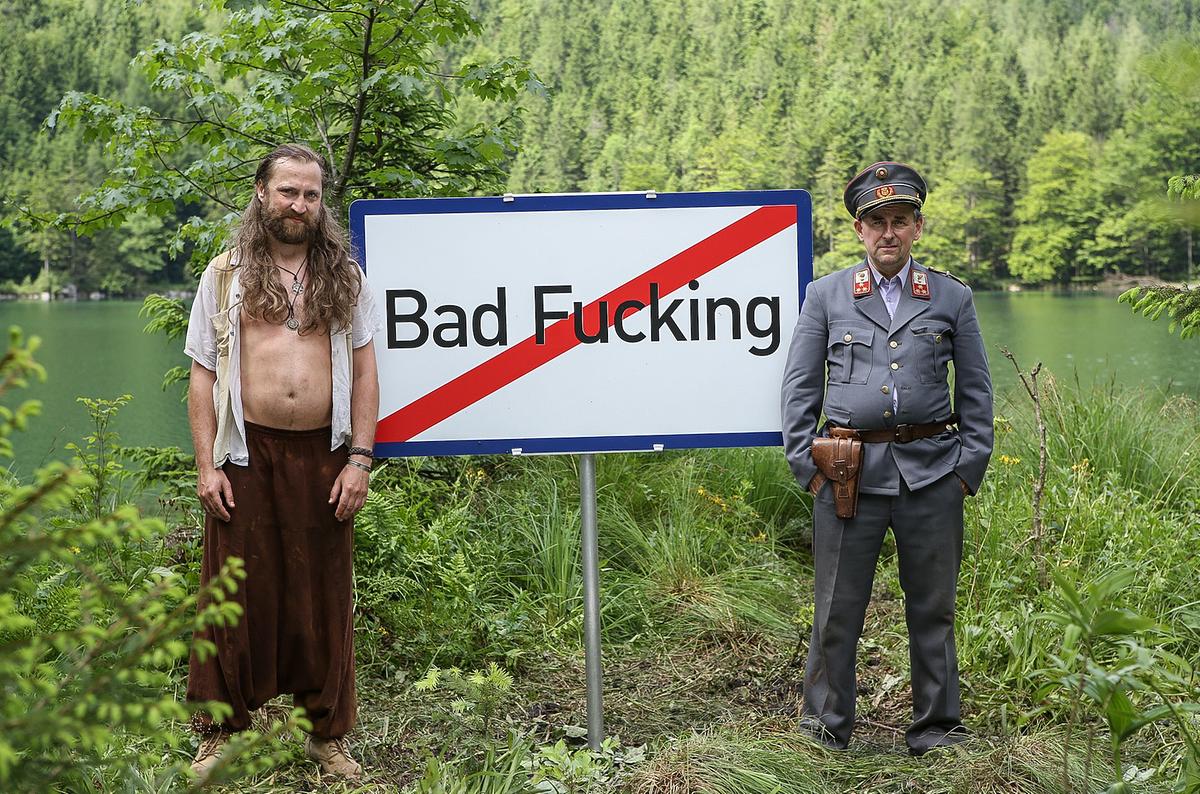 © ORF/MR Film/Petro Domenigg Bad Fucking; Im Bild: Christian Strasser mit offenem Hemd und langen Haaren und Johannes Silberschneider in Polizeiuniform stehen links und rechts des Ortsende-Schilds von Bad Fucking.