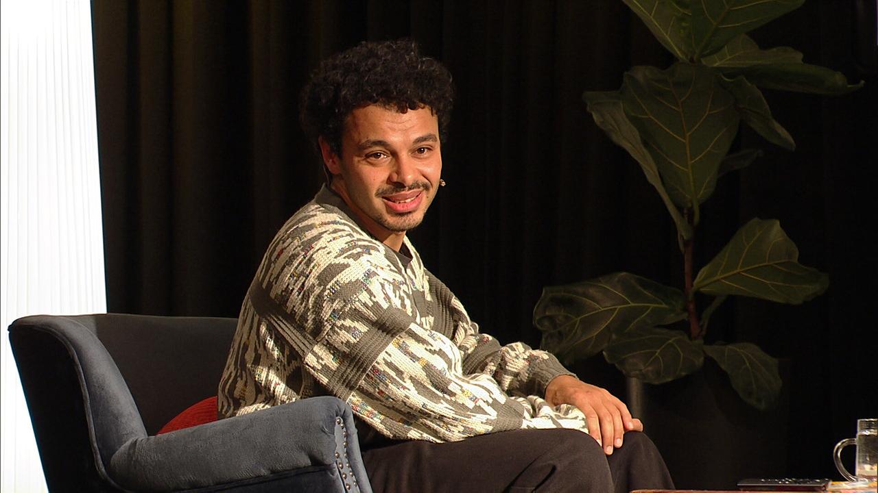 Romeo Kaltenbrunner sitzt in einem Sessel auf einer Bühne neben einer großen Zimmerpflanze.