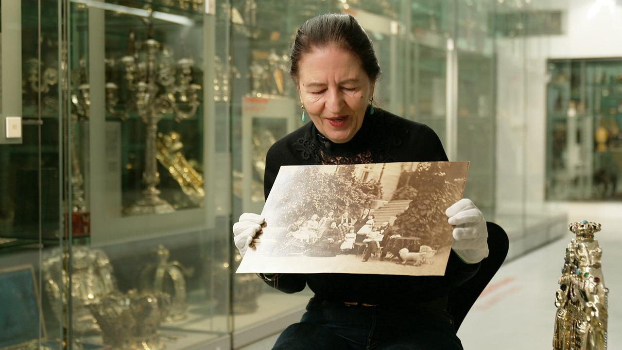 Gabriele Kohlbauer-Fritz zeigt mit weißen Handschuhen  in einem Ausstellungsraum ein altes schwarz‑weißes Gruppenfoto.