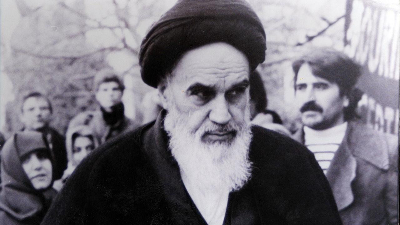 Schwarz-weiß-Aufnahme von Ayatollah Ruhollah Chomeini im Freien, im Hintergrund weitere Menschen.