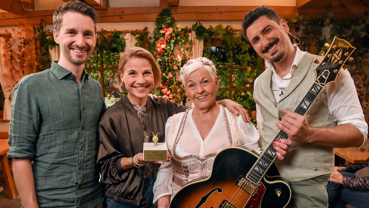 © ORF/Hubert Mican Clemens Maria Schreiner, Kristina Sprenger mit Trophäe in der Hand, Jazz Gitti und Lukas Maierhofer mit Gitarre posieren in rustikaler Kulisse beim ORF III Heurigenquiz.