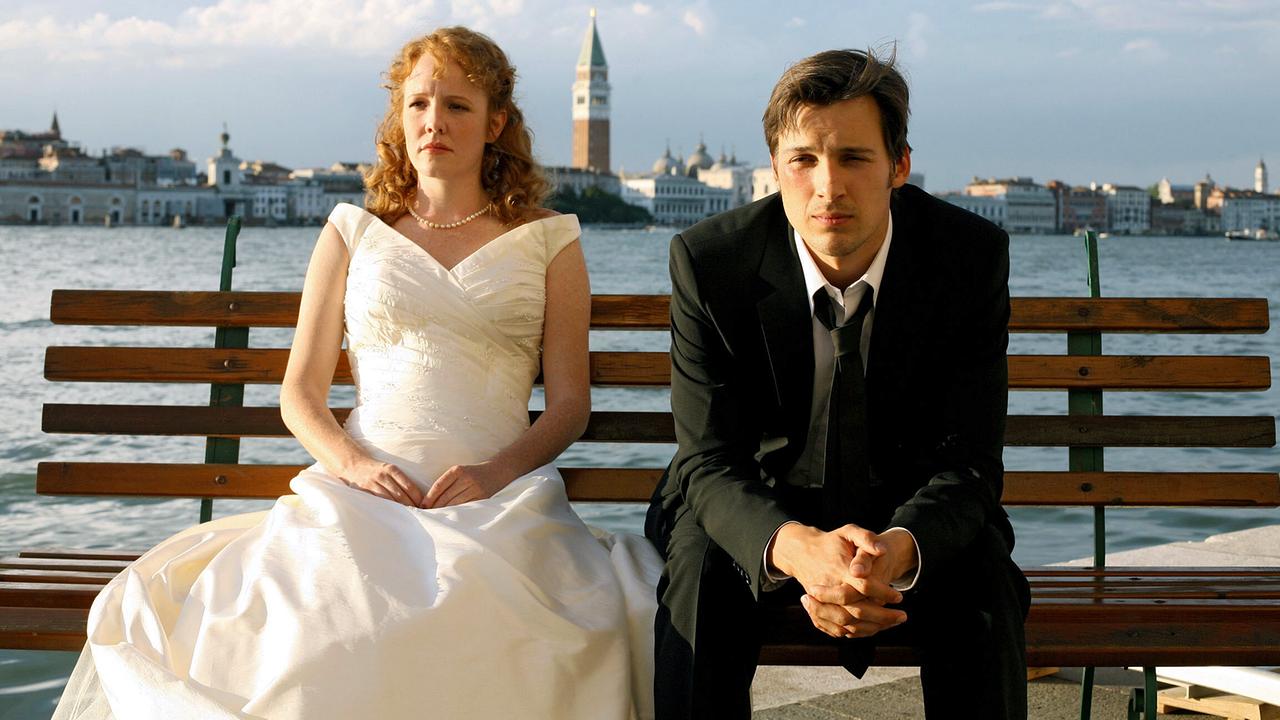 Anna und Max in Hochzeitskleidung sitzen auf einer Bank am Wasser vor der Kulisse von Venedig.