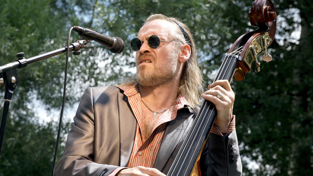 Johannes Krisch als Paul Schremser mit langen Haaren und Sonnenbrille spielt Kontrabass und singt in ein Mikrofon im Freien.