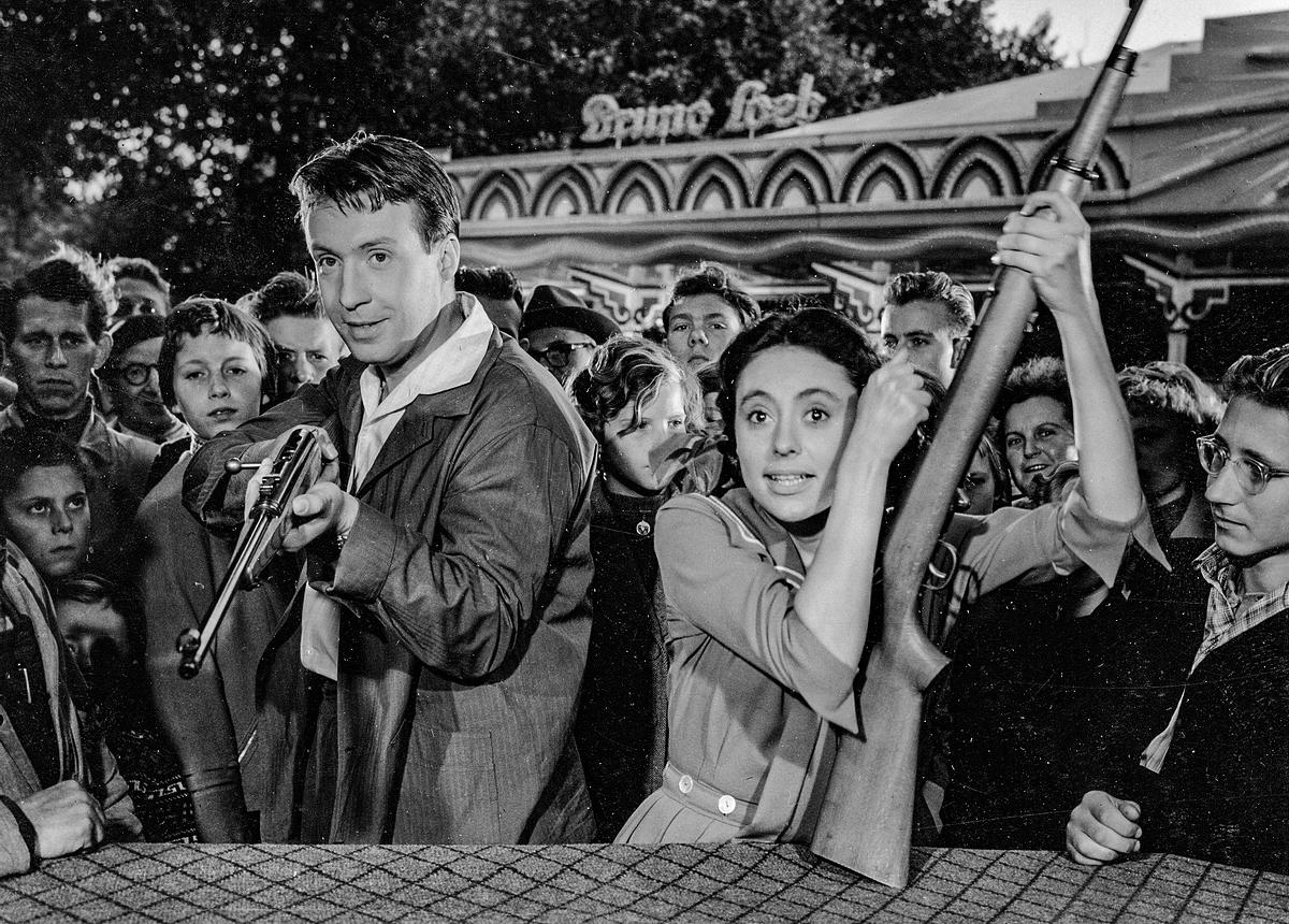 © ORF/CCC Film Liebe, Tanz und 1000 Schlager (Originaltitel: Mein Liebling Caterina (DEU 1955)); Im Bild: Peter Alexander und Caterina Valente vor einer Schießbude. Beide haben ein Gewehr in der Hand, Peter Alexander schießt, Caterina Valente lädt ihr Gewehr. Sie sind umringt von Menschen, die ihnen zusehen.