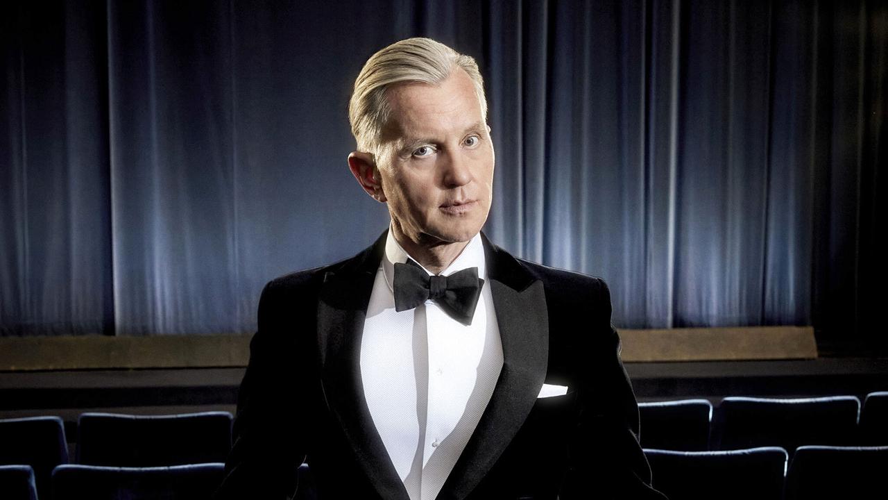 Max Raabe im Smoking steht in einem leeren Theatersaal und blickt in die Kamera.