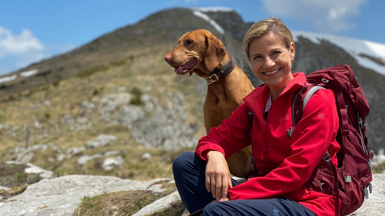 Kristina Sprenger -  mit Hund - sitz auf einem Stein. Sie hat einen roten Rucksack umgehängt und lächelt.
