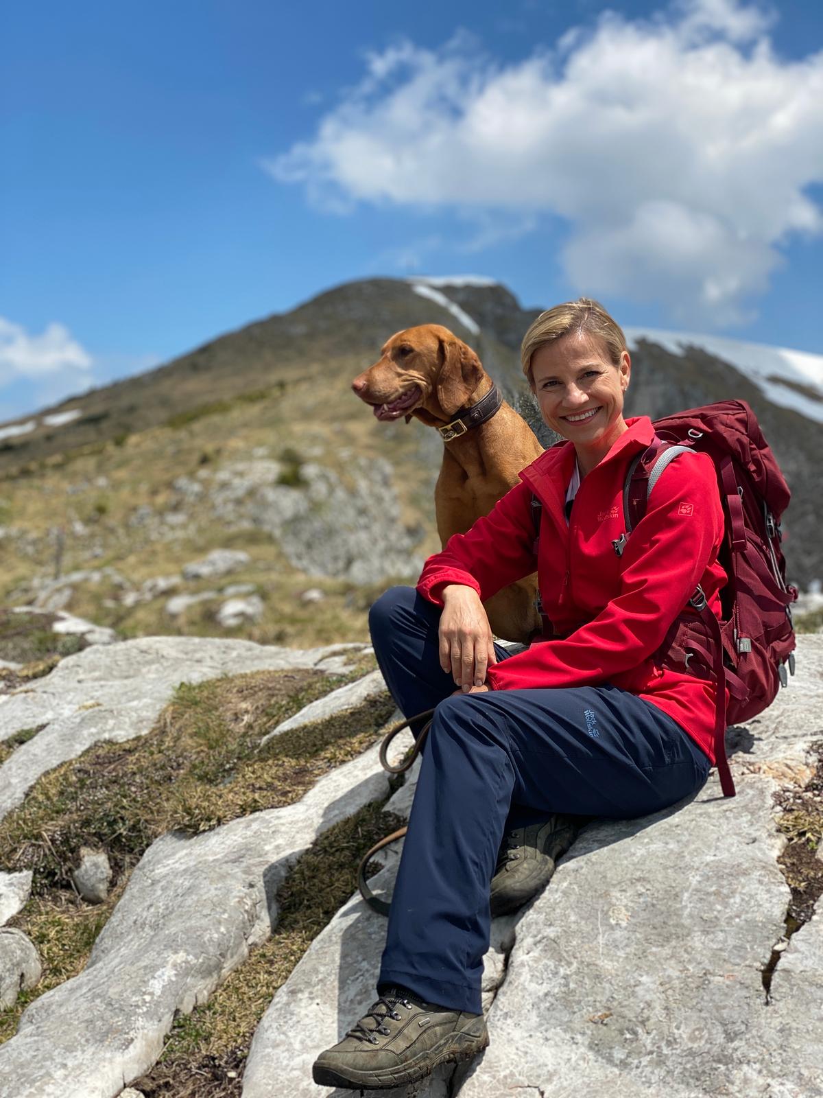 Kristina Sprenger -  mit Hund - sitz auf einem Stein. Sie hat einen roten Rucksack umgehängt und lächelt.