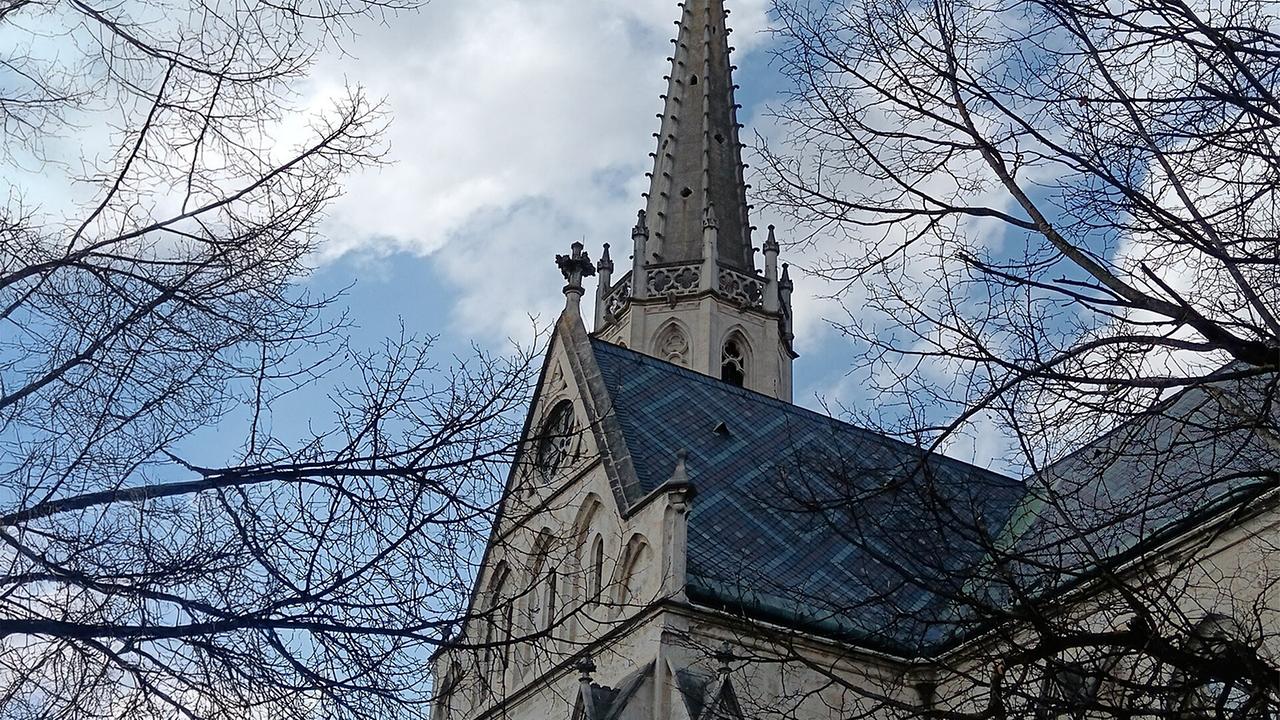 Der spitze Kirchturm und das Dach der gotischen Stadtpfarrkirche Bad Hall sind von kahlen Ästen eingerahmt vor einem teils bewölkten Himmel.