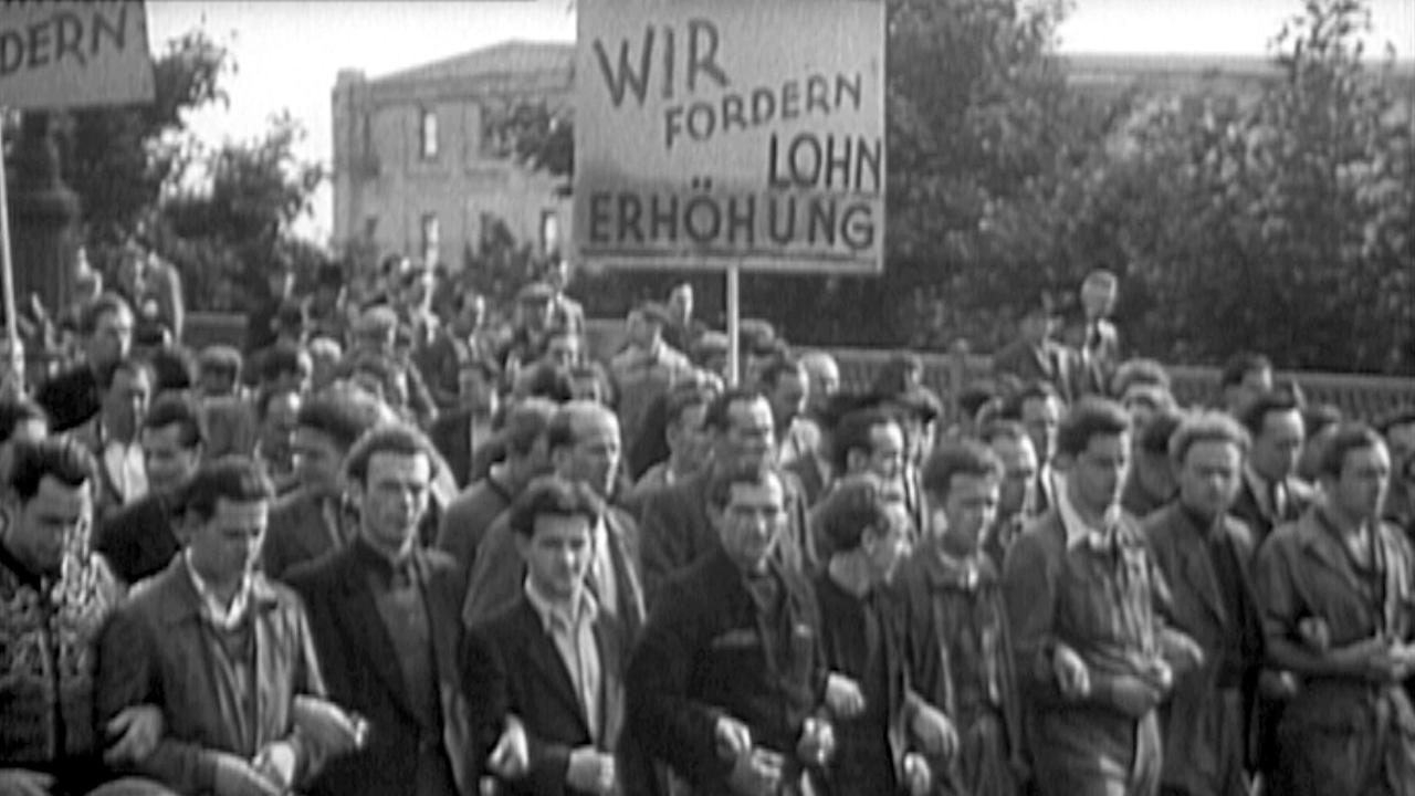 Streik in den 1950er-Jahren. Männer marschieren mit einem Schild auf dem steht: "Wir fordern Lohnerhöhung"