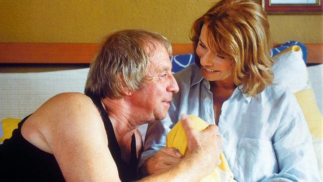 Ulrich Pleitgen und Senta Berger sitzen eng nebeneinander im Bett und lächeln sich an.