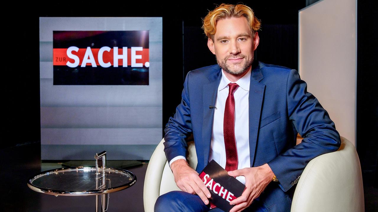 © ORF/Roman Zach-Kiesling "zur SACHE"-Moderator Reiner Reitsamer sitzt im Studio neben einem Glas-Couchtisch.