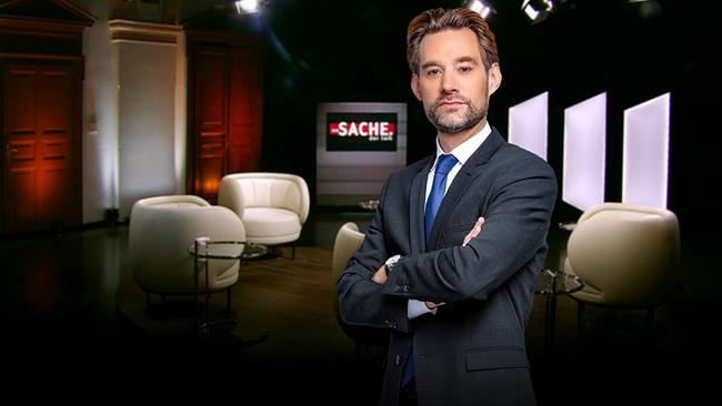 Reiner Reitsamer in Anzug und Krawatte steht mit verschränkten Armen im "zur SACHE"-Studio, mit weißen Sesseln und einem Bildschirm mit der Aufschrift „ZUR SACHE“ im Hintergrund