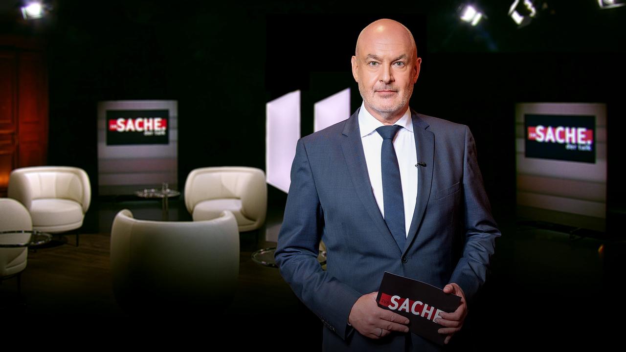 Wolfgang Geier in Anzug und Krawatte steht im "zur SACHE"-Studio, im Hintergrund weiße Coachsessel und Bildschirme mit "zur SACHE"-Aufschrift.