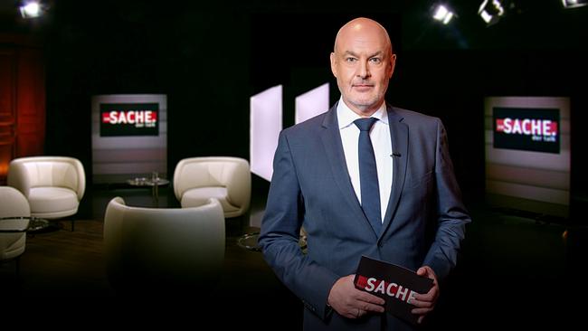 Wolfgang Geier in Anzug und Krawatte steht im "zur SACHE"-Studio, im Hintergrund weiße Coachsessel und Bildschirme mit "zur SACHE"-Aufschrift.